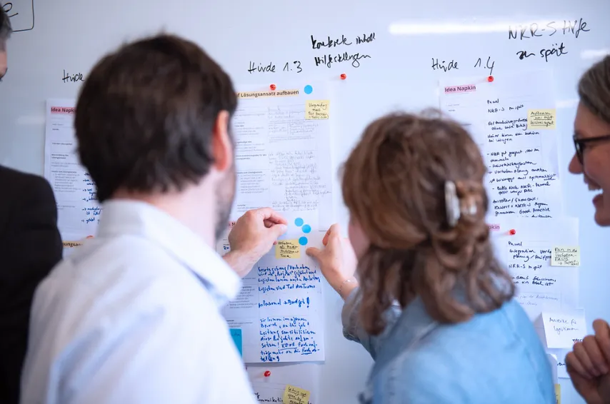 Mehrere Personen vor einem großen Whiteboard, an dem sie Klebepunkte an bereits vorhandenen Papieren und Post-its anbringen.