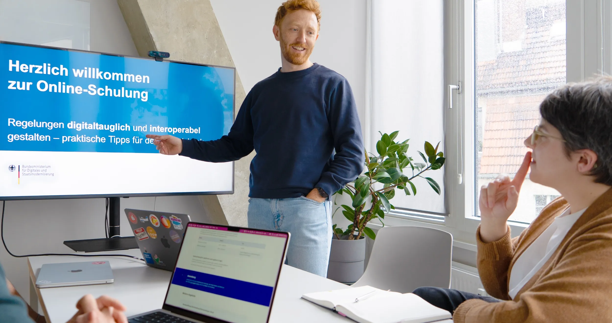 Ein Mann leitet eine Präsentation vor einem Bildschirm mit der Aufschrift „Herzlich willkommen zur Online-Schulung“ für Personen an einem Besprechungstisch mit Laptops.